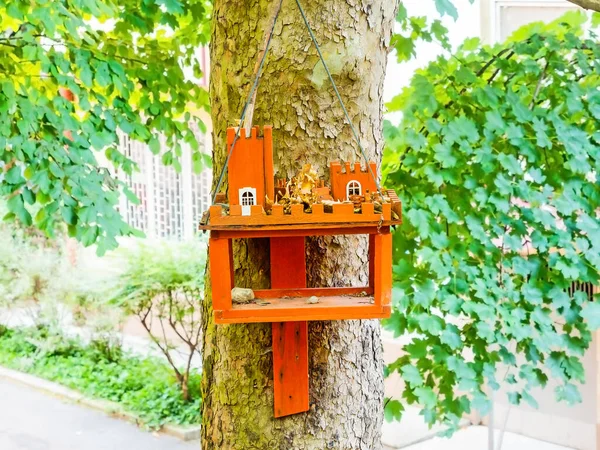 Orange wooden bird feeder with castle decor hanging on tree trunk with green foliage — Stock Photo, Image