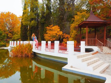 Kızıl saçlı kız, yeşil çamurlu bir gölün üzerindeki köprüde oturuyor. Parkta, sonbahar ağaçlarının arka planında ve bir çardakta.