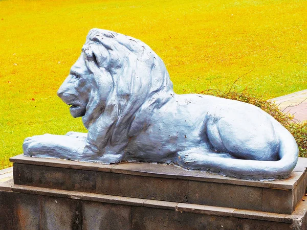Silver statue of a lion lying on a granite pedestal against the background of a yellow green lawn