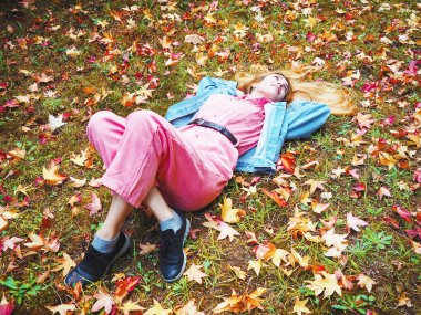 Pembe tulumlu bir kız sonbahar yapraklarıyla bezenmiş bir çayırda bacak bacak üstüne atmış yatıyor.