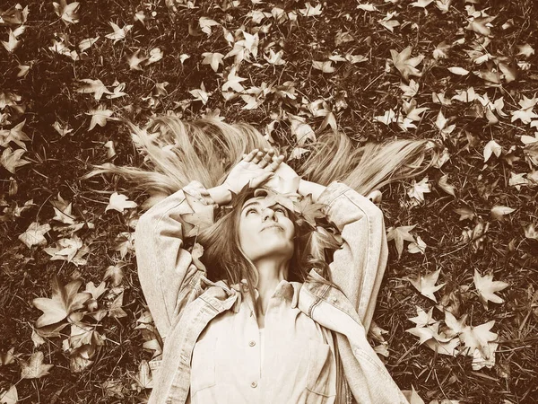 stock image A girl lies in a meadow covered with maple leaves with her palms folded above her head. Sepia filter