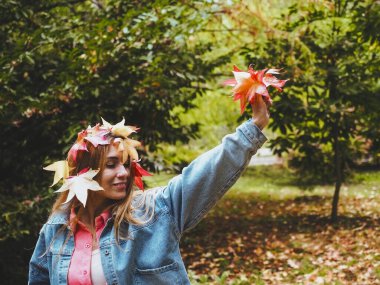Kırmızı akçaağaç yapraklarıyla süslenmiş gülen kız parkta elinde bir buket sonbahar yaprağı tutuyor.