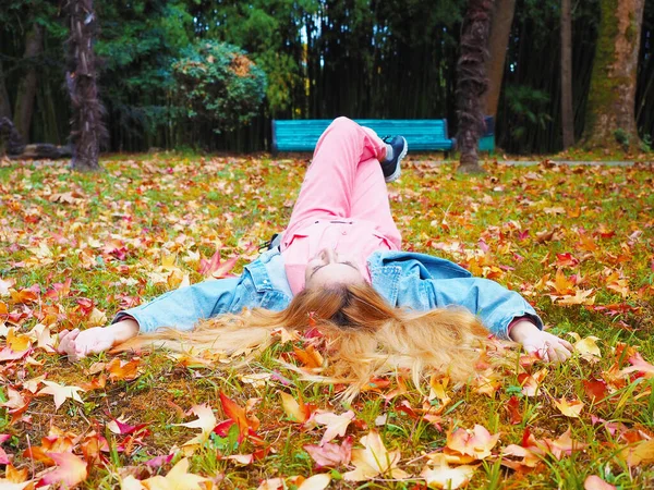 Η κοκκινομάλλα ξαπλώνει με σταυρωμένα τα πόδια σε ένα πάρκο σε ένα λιβάδι γεμάτο φθινοπωρινά φύλλα. — Φωτογραφία Αρχείου