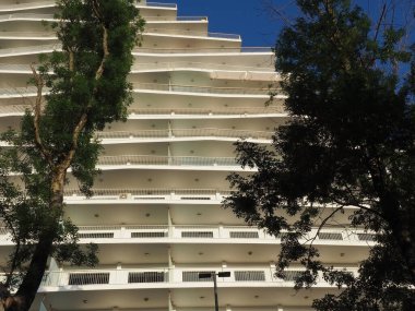 Önünde yeşil ağaçlar ve balkonları olan beyaz, modern bir binanın detayları. Fotoğraf aşağıdan yukarıya