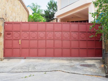 Kalesi olan kırmızı demir bir çit, geometrik desenler ve keskin örümcekler bir yerleşim binasını ve yeşil ağaçlarla çevrili bir avluyu kaplar.