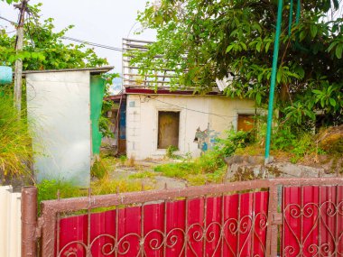 Eski, terk edilmiş ve çimenlerle kaplı, yıkık dökük çatılı bir ev ve metal ve kırmızı levhalardan yapılmış bir çitin arkasındaki kırık camlar.