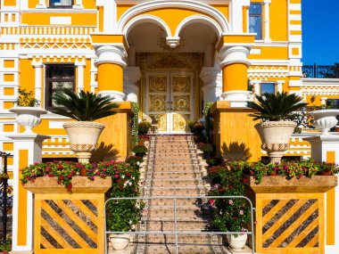 Beyaz süslemeli sarı bir bina parçası, sütunlar, altın desenli kapılar ve halıyla kaplı merdivenler.