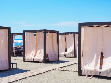 Kumsaldaki bungalovlar, açık güneşli bir günde mavi gökyüzünün altında aralarında ahşap patikalar olan kumların üzerinde dururlar.