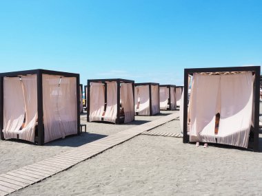 Açık güneşli bir günde, açık bir gökyüzünün altında, kumların üzerinde, aralarında ahşap patikalar bulunan, ışık perdeli plaj kamaraları sıralanır.