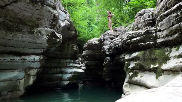 Girl jumping from a steep mossy canyon into a deep turquoise river — Stock Video