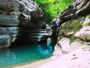 Parlak turkuaz sakin bir nehir, tepesinde parlak yeşil ağaçlar olan yosunlu kayaların arasından akar. Kanyon beyaz kayalıkları
