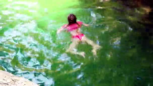 Una chica en un traje de baño rosa nada a lo largo de un río verde brillante soleado en un desfiladero entre las rocas — Vídeo de stock