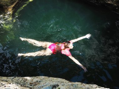 Pembe mayo giymiş bir kız, kolları ve bacaklarıyla nehirde yüzüyor. Kayaların arasında güneşin altında parıldıyor.