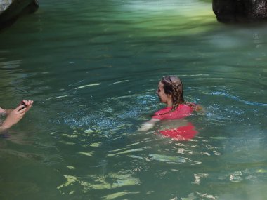 Kameraya poz veren bir kız kayalık bir vadide turkuaz bir nehirde yüzüyor.