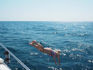 Bir kız açık mavi denize bir yattan atlıyor. Güneşin aydınlattığı açık bir günde.