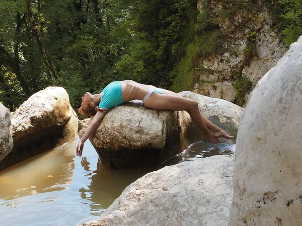 A girl in a blue swimsuit lies on a large stone in the middle of muddy water, rocks and surrounded by a green grove —  Fotos de Stock