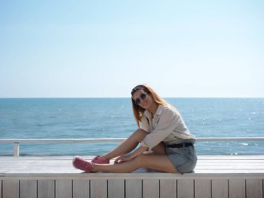 Gülümseyen kız, ahşap bir sınırda oturuyor denizin arka planında ve berrak mavi gökyüzünde