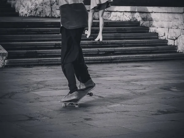 De man rijdt op een skateboard en bereidt zich voor op een stunt tegen de achtergrond van een meisje dat de trap op klimt in een minirok. Zwart-wit foto — Stockfoto