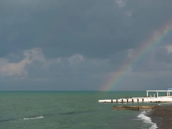 Multicolored rainbow in gray cloudy sky over turquoise sea and white pier Royalty Free Stock Images