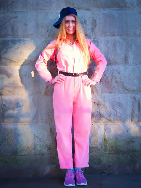 Smiling girl in pink jumpsuit stands by a stone wall in sunlight — Stock Photo, Image