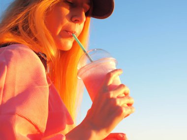 Kızıl saçlı kız mavi gökyüzünün arka planındaki bir tüpten milkshake içiyor.