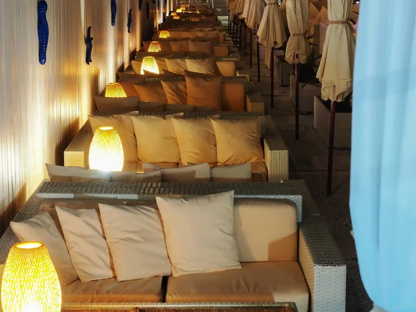 Φως καναπέδες μπαστούνι με μαξιλάρια και λάμπες σταθεί σε μια σειρά στο ημι-σκοτάδι. Πολυτελής χώρος αναψυχής παραλίας. Παραλία καφέ — Φωτογραφία Αρχείου