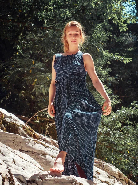 Ένα κορίτσι με μακρύ φόρεμα περπατάει ξυπόλητο σε μια βραχώδη περιοχή κάτω από πράσινα δέντρα που φωτίζονται από τον καλοκαιρινό ήλιο.. — Φωτογραφία Αρχείου