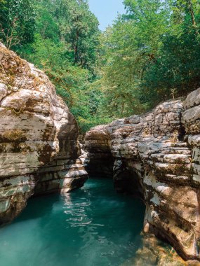 Zengin turkuaz suyu olan sakin bir nehir, tepesinde yaprak döken ağaçlarla dolu bir vadiye akar.