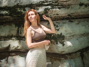 Bir kız güneşli bir yaz gününde yosun kaplı kayanın yanında durur.