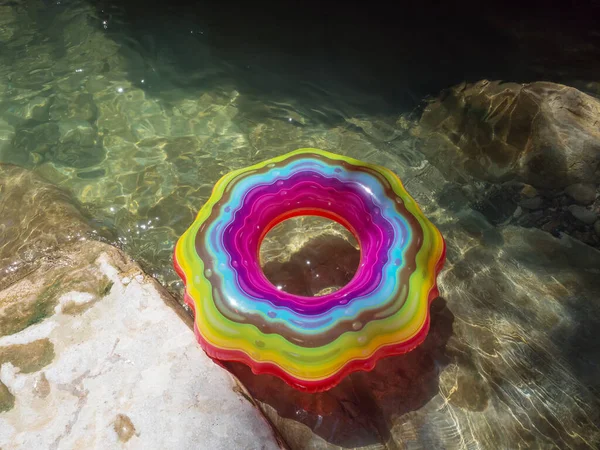 Círculo de natación con patrones multicolores en un río claro con un fondo rocoso bajo los rayos del sol —  Fotos de Stock