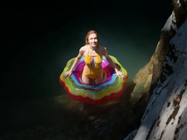 Chica sonriente en un círculo de natación multicolor se encuentra en el agua en los rayos del sol en un barranco sombreado de cañones —  Fotos de Stock