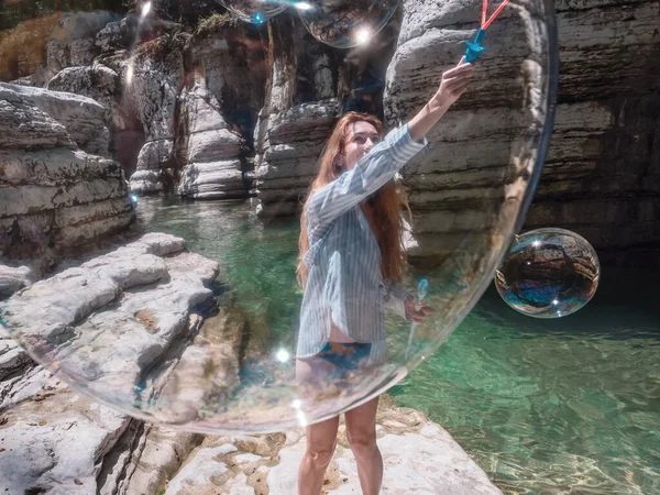 La chica sopla enormes burbujas de jabón en la garganta del cañón cerca del río claro. Vista a través de la burbuja —  Fotos de Stock