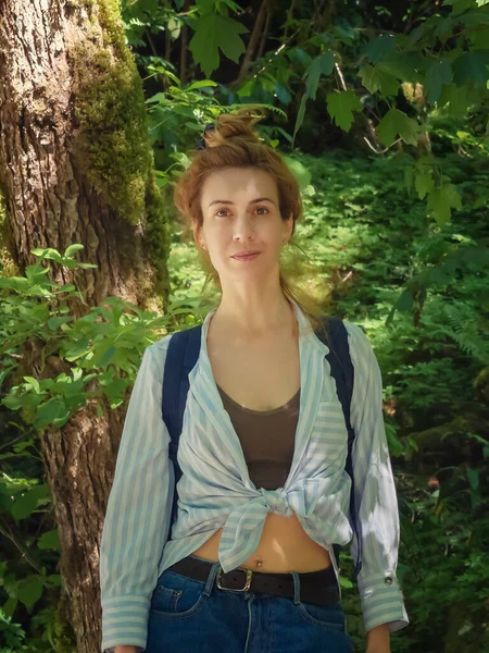 Улыбающаяся девушка в вязаной рубашке стоит на фоне густой зеленой растительности в лесу — стоковое фото
