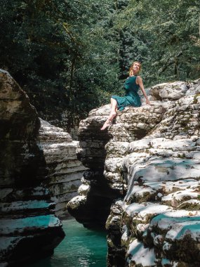 Turkuaz suyun üzerindeki bir kanyonda, yaprak döken ağaçların altında bir kız oturuyor. Filtreli fotoğraf