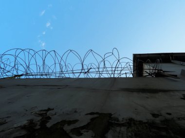 Mavi gökyüzüne karşı kabuk soyulmuş alçı ve dikenli tel ile yüksek beton çitler. Alt görünüm