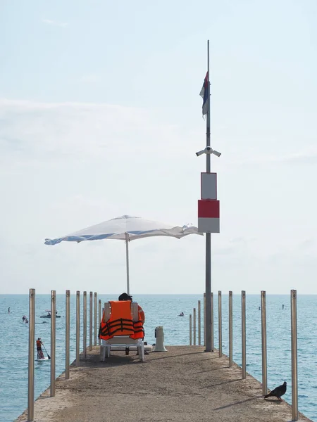 Salva-vidas senta-se em uma espreguiçadeira no cais sob um guarda-chuva junto ao mar em um dia ensolarado de verão — Fotografia de Stock