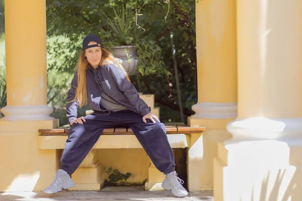 Una chica con un traje de pana hip-hop se sienta en un banco entre las columnas del parque — Foto de Stock