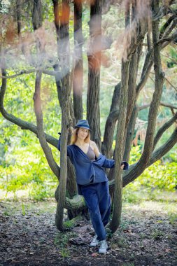 Mavi elbiseli ve şapkalı bir kız parktaki dallı bir ağacın gövdesine tutunuyor.