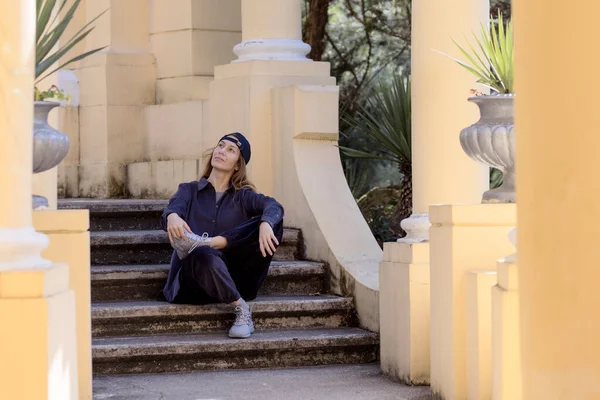 Женщина в хип-хоп стиле сидит на ступеньках среди колонн в парке — стоковое фото