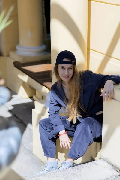 Sonriente rubia de pelo largo vestida de estilo deportivo se sienta rodeada de la vieja arquitectura del parque en un día soleado — Foto de Stock