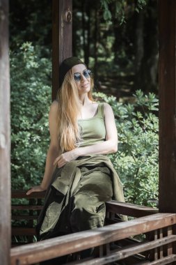 Hip-hop kıyafetli, uzun saçlı, gülümseyen bir kadın yeşil bir yaz parkındaki kirişe yaslanmış bir çardağın ahşap korkuluğunda oturur..