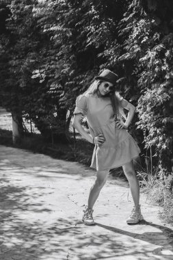 Yazlık elbiseli ve şapkalı bir kız parkta ayak parmakları içe dönük dururken ara sokakta poz veriyor. Siyah beyaz fotoğraf