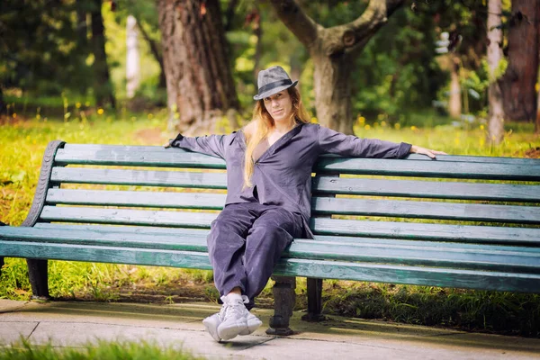 Uma mulher de cabelos longos com roupas lilás e um chapéu senta-se em um banco de parque. Dançarina de rua. Vagabundo — Fotografia de Stock