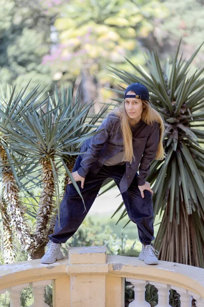 Una chica con una gorra con una visera en la espalda y un traje de velour hip-hop está en una pose en la acera en el parque cerca de las palmeras — Foto de Stock
