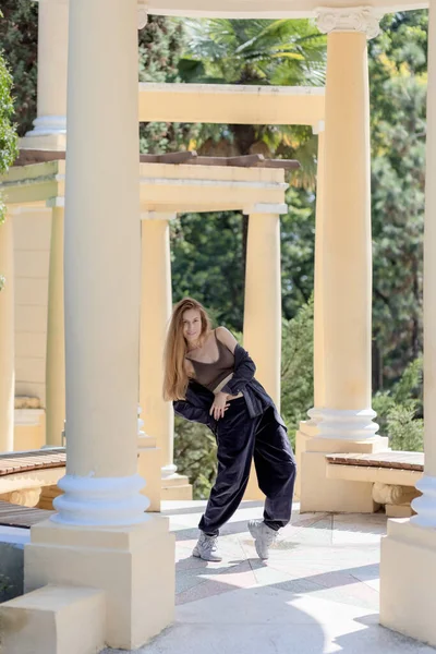 A long-haired woman in wide corduroy pants and a shirt dropped from her shoulders stands in a curved position among the columns in the park