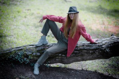 Sokak kıyafeti giymiş uzun saçlı bir kız ve parktaki devrilmiş bir ağacın gövdesinde oturan bir şapka.