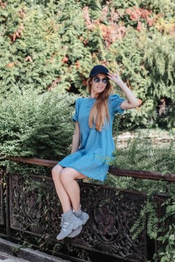 Mavi yaz elbisesi içinde gülümseyen kız yeşil bir parkta metal bir korkulukta oturuyor.