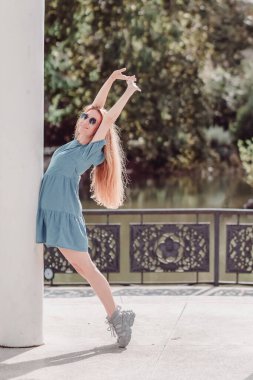 Kısa elbiseli, gülümseyen bir kız ayak parmaklarının ucunda duran bir sütuna yaslanıyor ve ellerini parktaki yeşil bir göletin arka planına doğru kaldırıyor.
