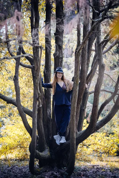 Усміхнена дівчина в капелюсі і синій повсякденний одяг стоїть в центрі гіллястого дерева в осінньому парку — стокове фото