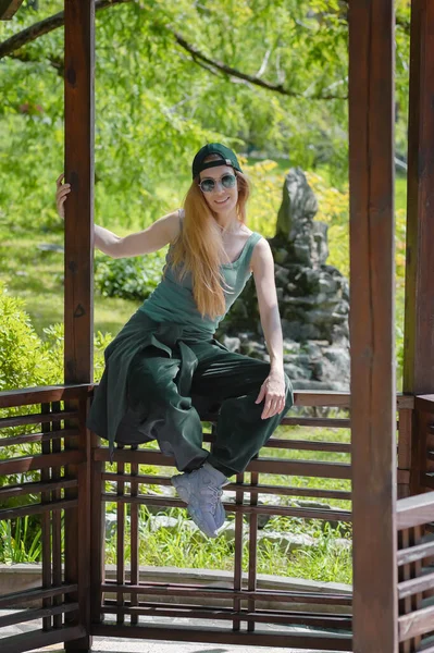 Mulher sorridente em roupa de hip-hop esmeralda senta-se ao lado de um mirante de madeira em um parque verde de verão — Fotografia de Stock